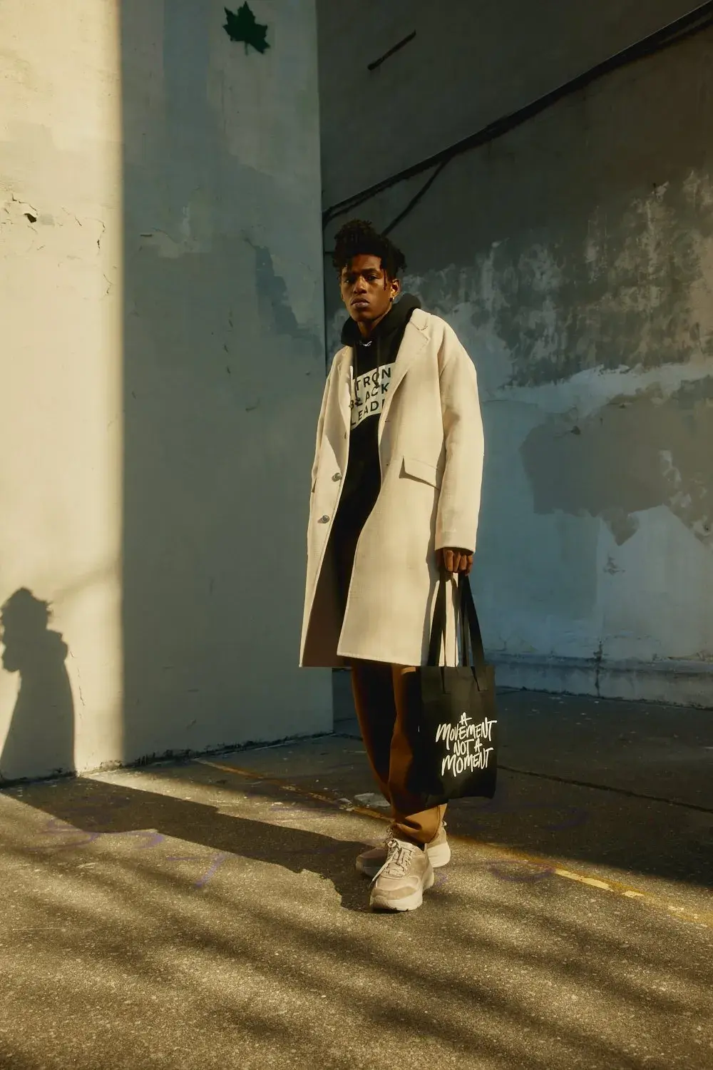 A person wears a light coat over a Strong Black Lead sweatshirt and carries a black tote reading “A Movement Not A Moment” beside a sunlit concrete wall.