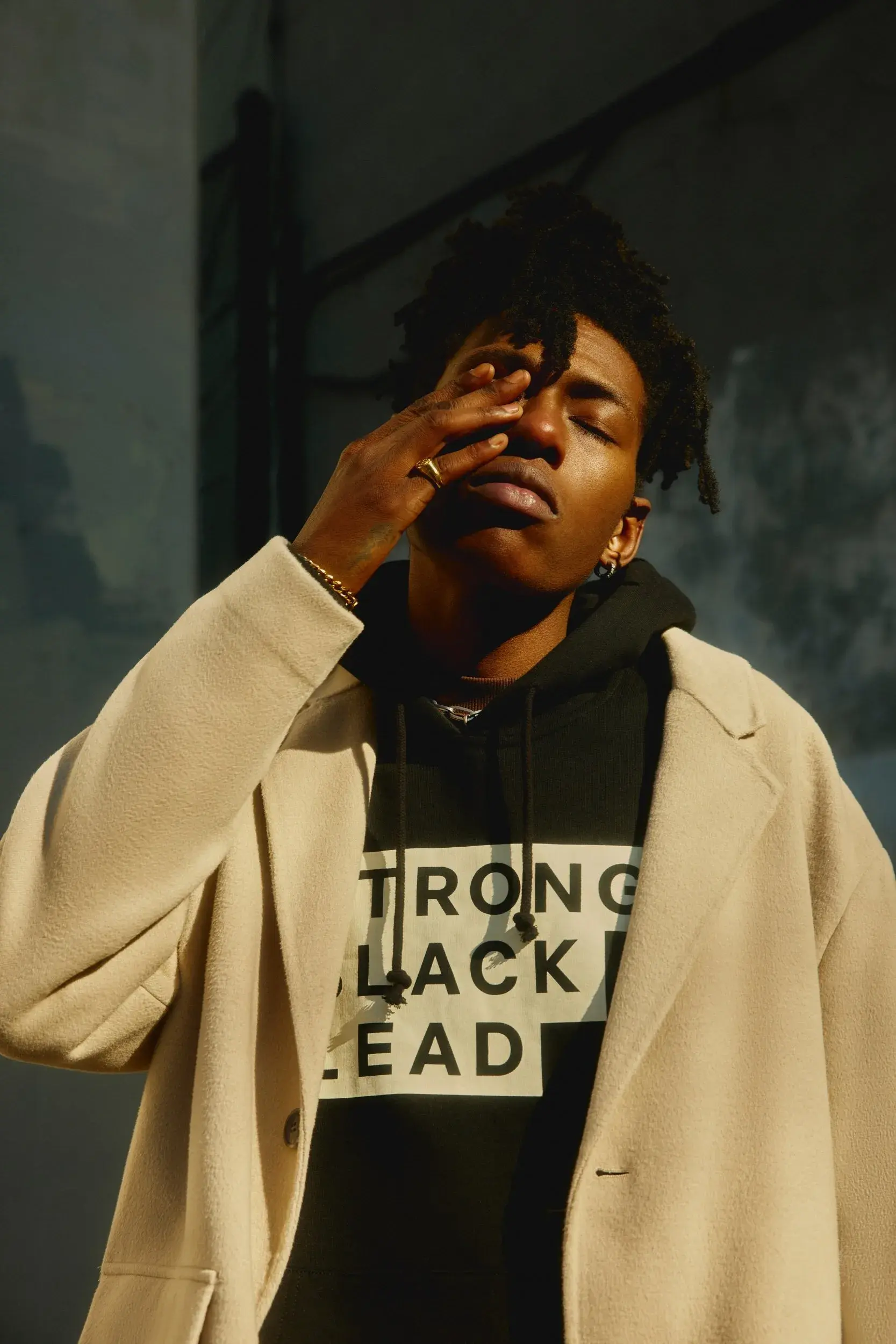 A person in a light coat wears a black hoodie featuring the stepped Strong Black Lead logo in a sunlit portrait.