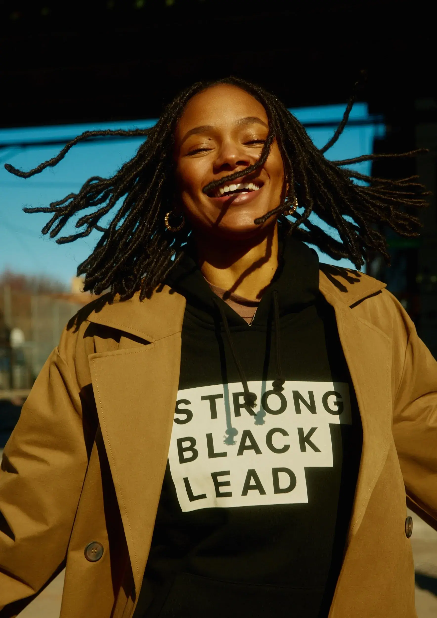 A smiling woman outdoors wears a tan coat over a black hoodie featuring the stacked Strong Black Lead wordmark.