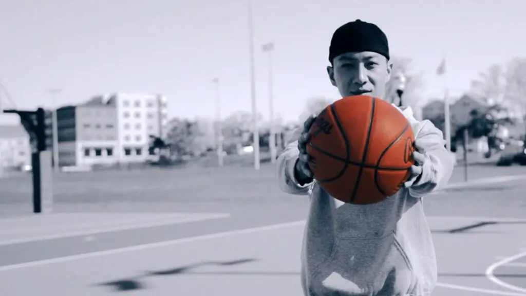 A young man holds an orange basketball toward the camera on an outdoor court, with the rest of the scene rendered in black and white.