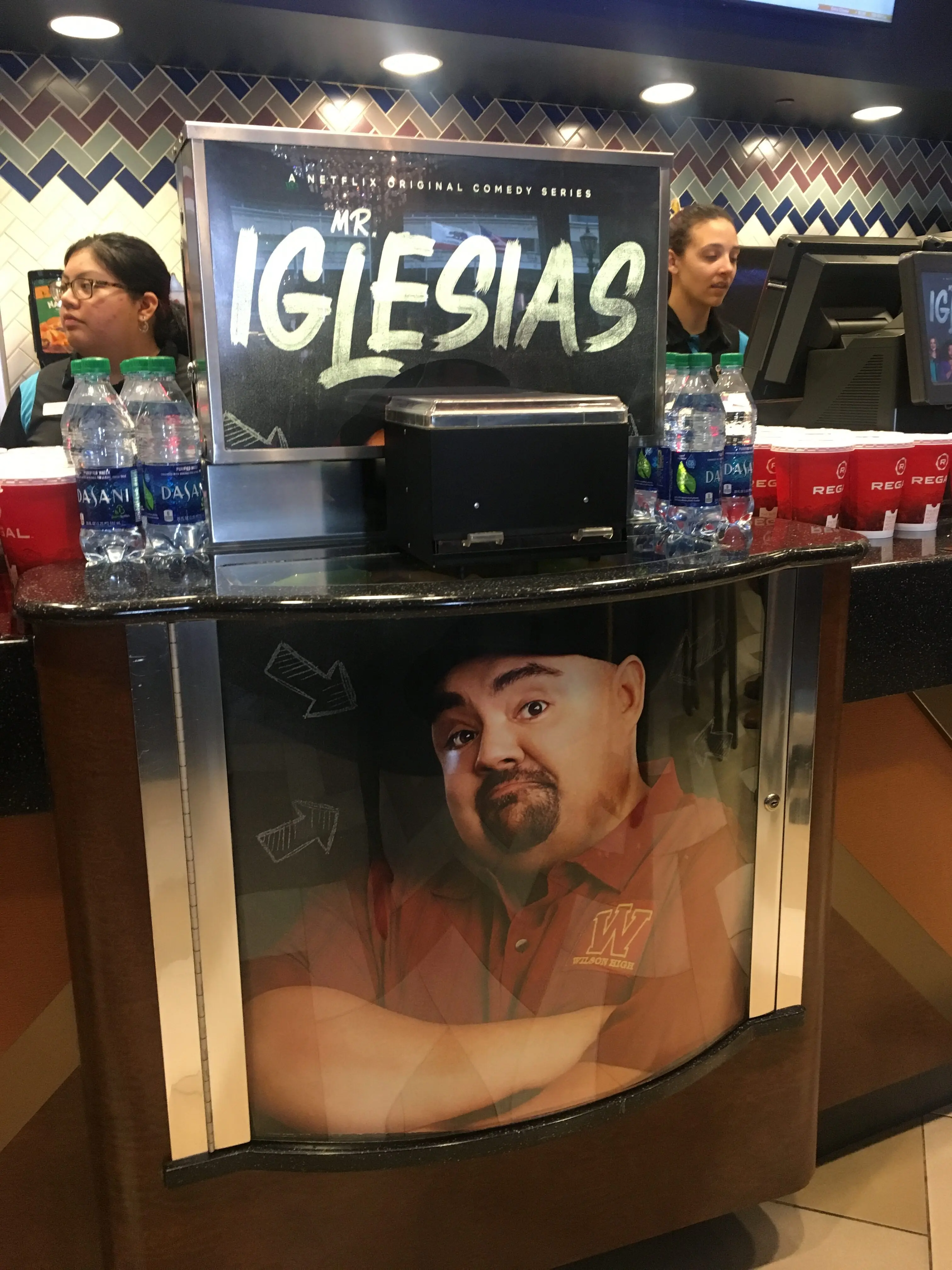 Theater concession counter branded with a chalk-style 'Mr. Iglesias' sign above a curved panel showing a man's portrait, two staff at registers, bottled water, and concession cups.