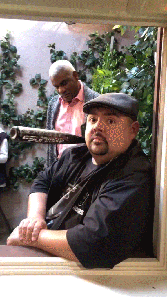 Two men framed in a vertical window crop: a man in a flat cap stares at the camera while a second man behind him balances a baseball bat on his shoulder against a leafy backdrop.