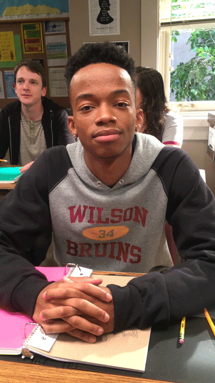Young Black student in a gray 'Wilson Bruins' hoodie sits at a classroom desk with hands folded on a notebook, looking at the camera with a neutral, slightly skeptical expression.