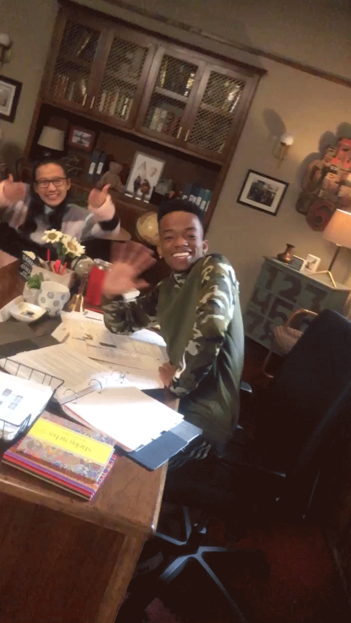 Two smiling students wave toward the camera while seated at a cluttered desk with binders, papers, a lamp, and books in a warmly lit classroom-like room in a vertical GIF loop.