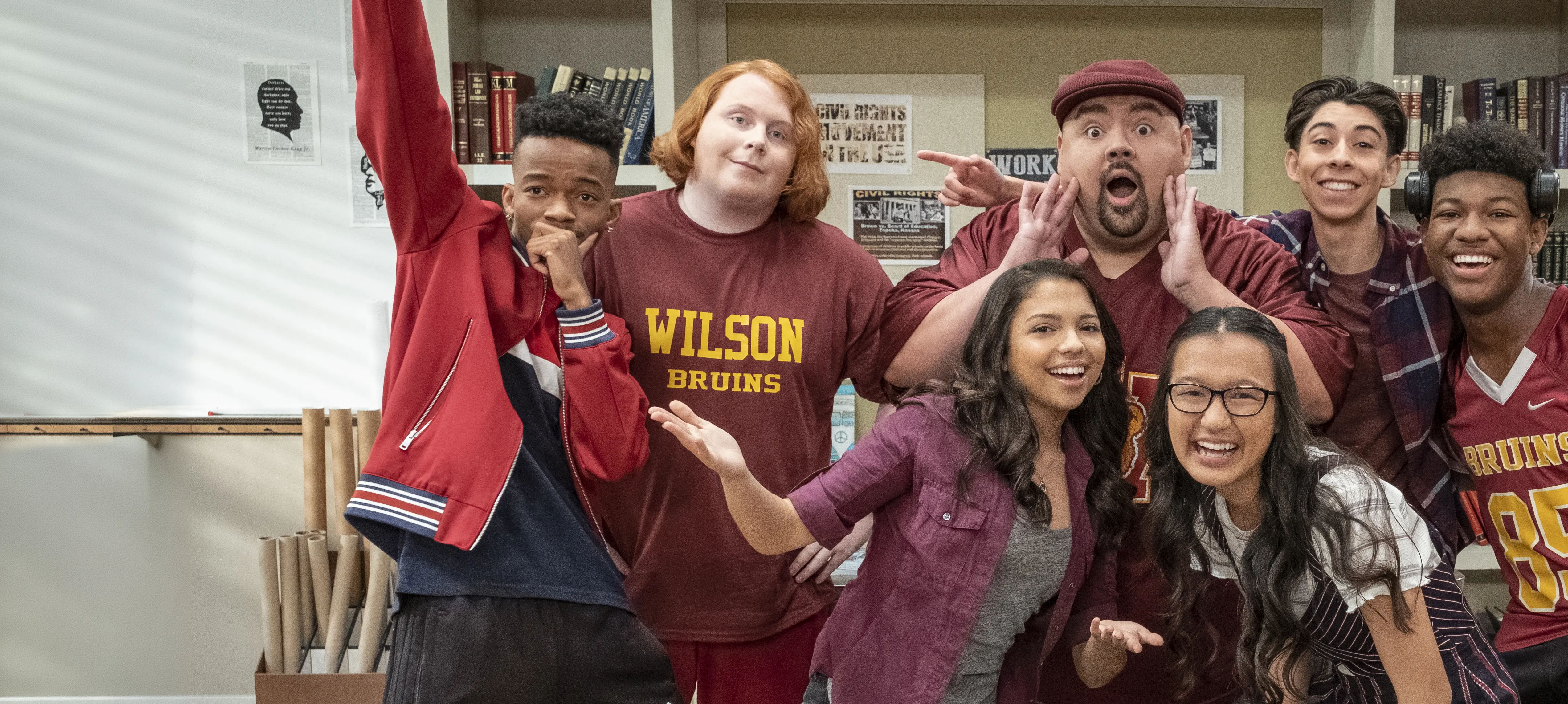 Seven high-school students and their teacher in a classroom pose for a playful group photo, several wearing maroon 'Wilson Bruins' shirts and one man in a maroon cap making an exaggerated surprised face.