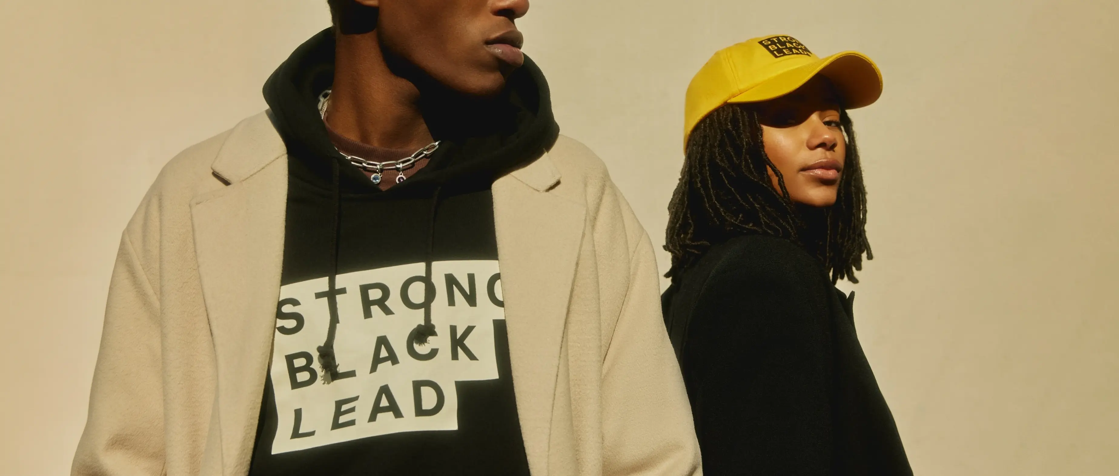 Two models wear Strong Black Lead apparel, including a black hoodie with the stepped wordmark and a yellow cap, against a warm neutral backdrop.