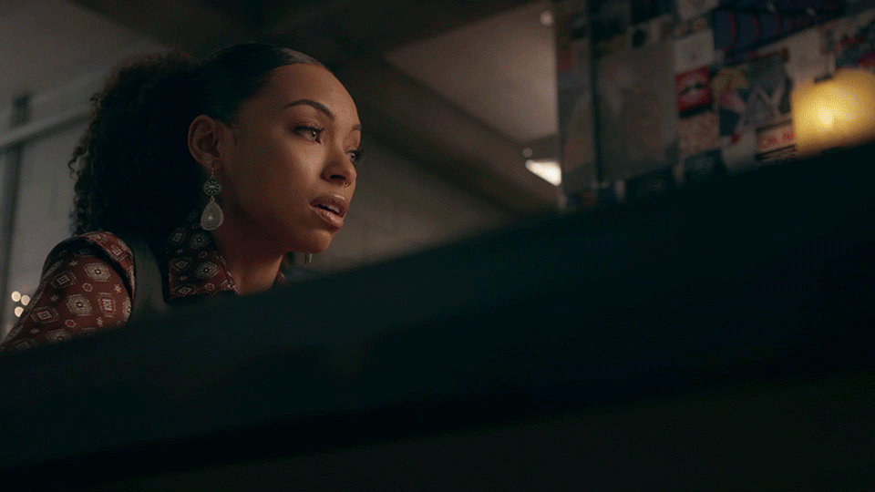 A woman in patterned clothing and dangling earrings looks toward an unseen screen in a dim room with photos and a glowing lamp behind her.