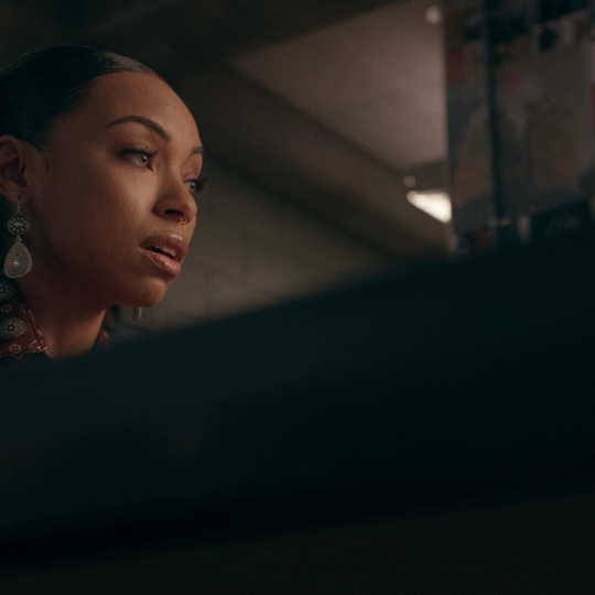 Close-up of a woman with pulled-back hair and dangling earrings looking tearful in a dimly lit interior.