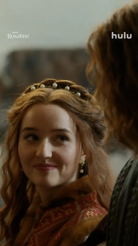 Rosaline, wearing a pearl-studded headpiece and patterned period dress, looks up with a knowing smile at a partially seen figure beside her.